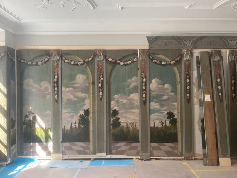 large room with decorative wall paintings, wooden pillars, checkered floor, unfinished ceiling
