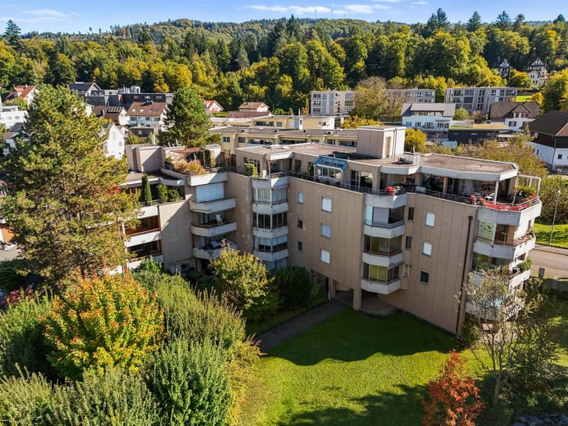 A multi-story building with balconies and terraces, surrounded by trees and grass.