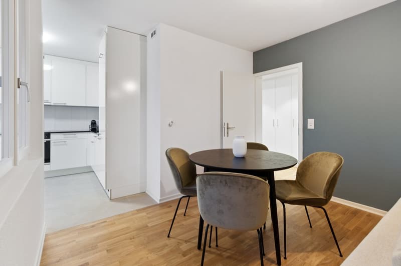 Open-concept living area with a round dining table and velvet chairs, hardwood floors, white walls, and a kitchen visible in the background with a dishwasher