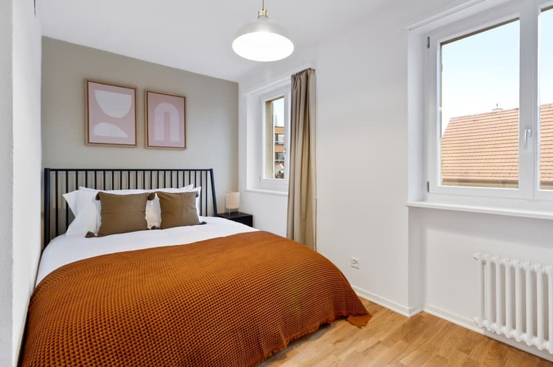 Spacious bedroom with hardwood floors, white walls, large window, black metal bed frame, orange bedspread, and two framed abstract art pieces on the wall.