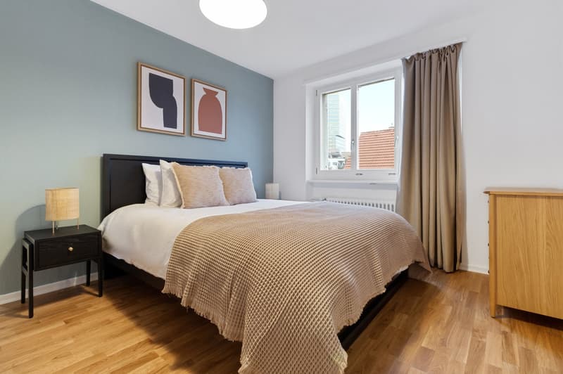 The image shows a cozy bedroom with a large bed, a black wooden headboard, and a beige patterned blanket. The walls are painted in a light blue-gray color, and there are two framed abstract art pieces hanging on the wall. The room has a large window that