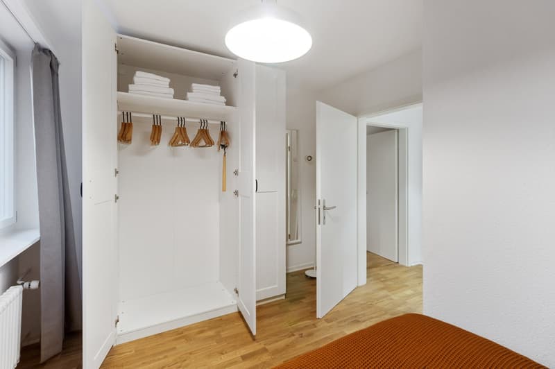 The image shows a bedroom with a built-in wardrobe. The wardrobe has shelves and hanging rods for storing clothes and linens. The room has hardwood floors and a window, and the walls are painted a light color. There is also a door leading to another room.