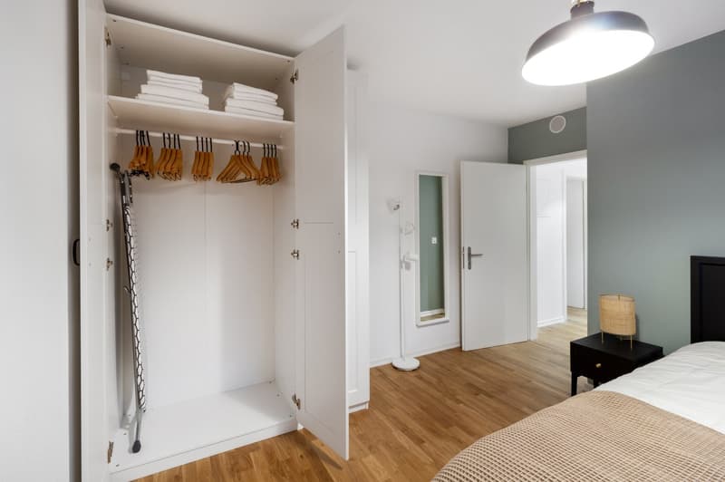Spacious bedroom with a built-in wardrobe, wooden flooring, and a private washing machine. The room has a clean and minimalist design with white walls and a neutral color scheme.