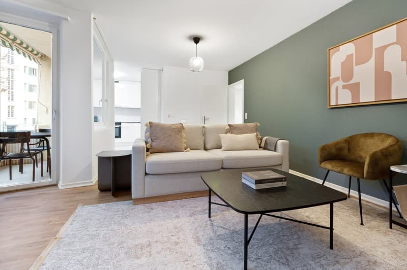 Spacious living room with large windows, light-colored sofa, black coffee table, armchair, and abstract artwork on the wall.