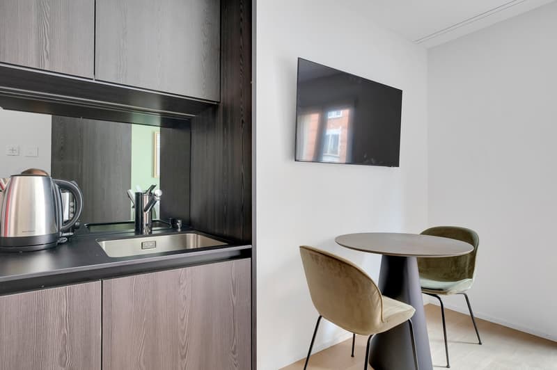 Modern kitchen with dark wood cabinets, stainless steel appliances including a dishwasher, and a small dining area with a round table and velvet chairs.