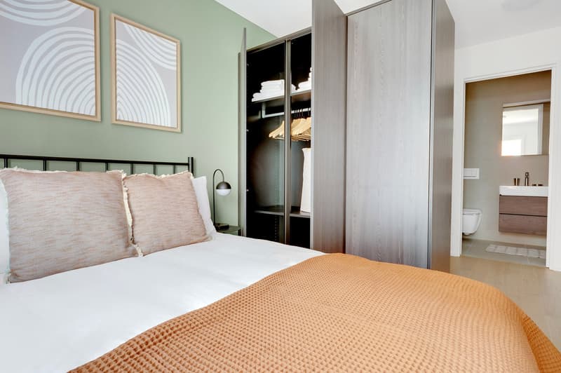 Spacious bedroom with a large bed, white bedding, and a plush orange blanket. The room features modern artwork on the walls, a built-in closet, and an ensuite bathroom visible through the open doorway.