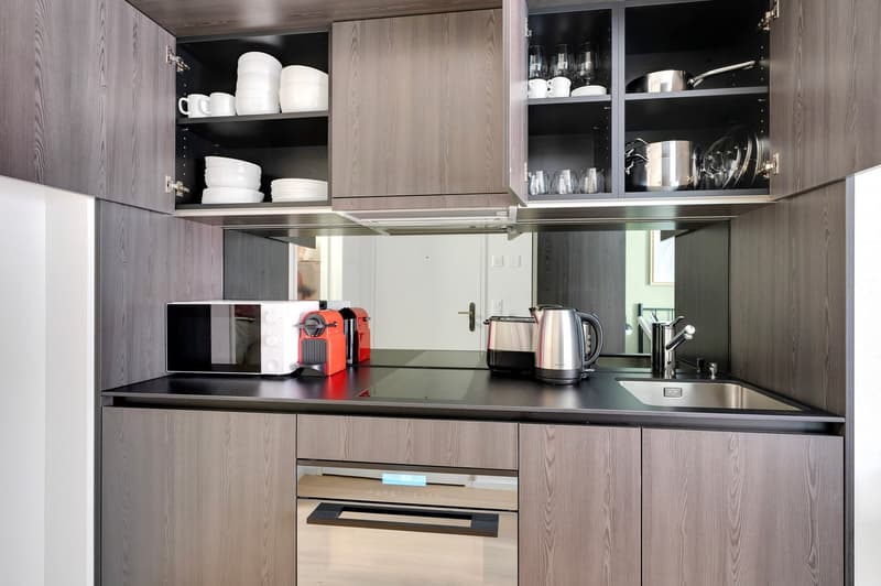 Modern kitchen with gray wood cabinets, black countertop, built-in appliances including a dishwasher and coffee maker, and open shelving displaying dishes and glassware.