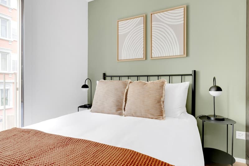 Spacious bedroom with white walls, large windows, black metal bed frame, white bedding, beige throw pillows, and abstract art prints on the wall. The room also features a small side table with a lamp.