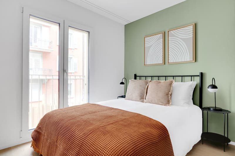 Spacious bedroom with large windows, light green walls, black metal bed frame, white bedding, orange knit blanket, and framed abstract art on the walls.