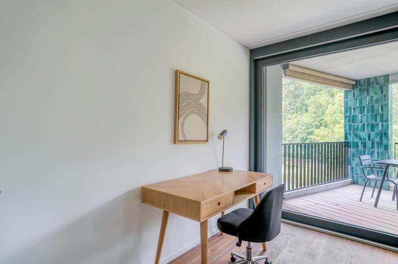 This image shows a modern and minimalist room with a wooden desk, a black chair, and a framed artwork on the wall. The room has large windows that provide a view of a balcony or terrace overlooking a wooded area.
