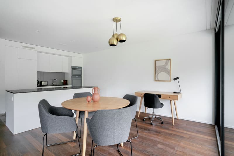The image shows an open-plan living area with a kitchen, dining table, and workspace. The kitchen features white cabinets and a gray countertop. The dining table has a round wooden top and is surrounded by gray upholstered chairs. The workspace has a wood