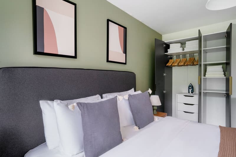 Spacious bedroom with a grey upholstered headboard, white bedding, and grey throw pillows. Two framed abstract art pieces hang on the wall. A wardrobe with shelves and hanging space is visible on the right side of the room.