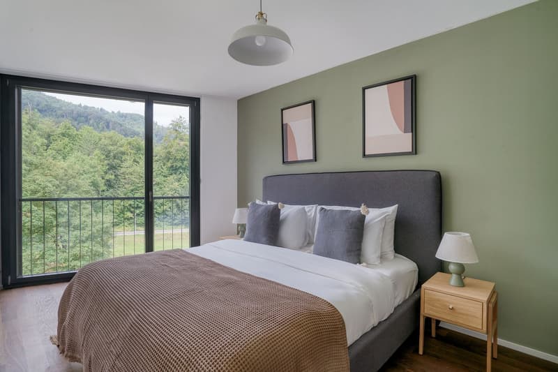 Spacious bedroom with large windows overlooking a forested landscape, grey upholstered bed frame, wooden nightstand, abstract artwork on the walls, and a pendant light fixture.