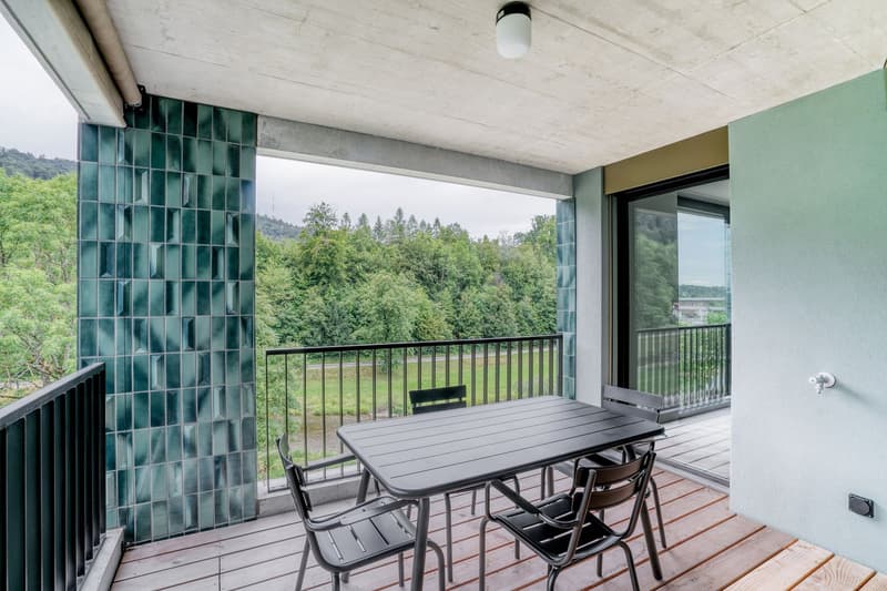 Covered balcony with glass block wall, wooden deck, outdoor dining table and chairs, overlooking a forested area