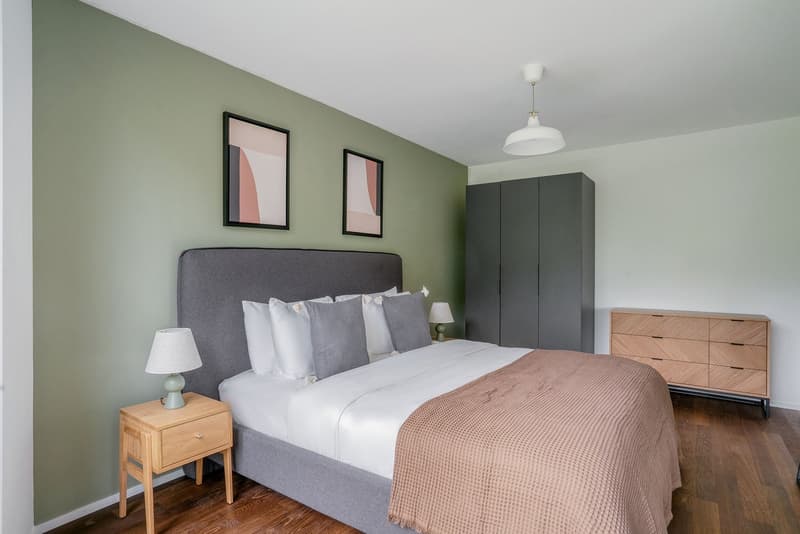 Spacious bedroom with green walls, gray upholstered bed frame, white bedding, wooden nightstands, and a large wardrobe. The room also features abstract artwork on the walls and a pendant light fixture.