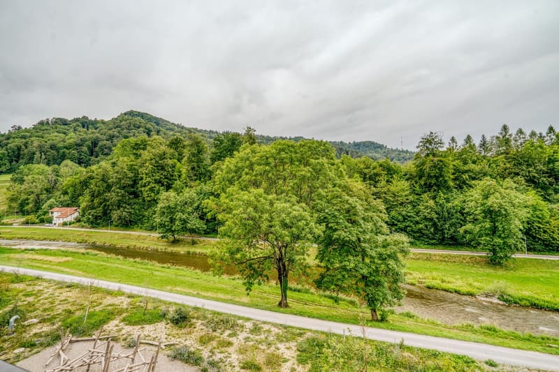 The image shows a scenic rural landscape with lush green trees and vegetation. There is a winding road running through the middle of the frame, with a small body of water visible in the distance. The landscape is surrounded by rolling hills and mountains,