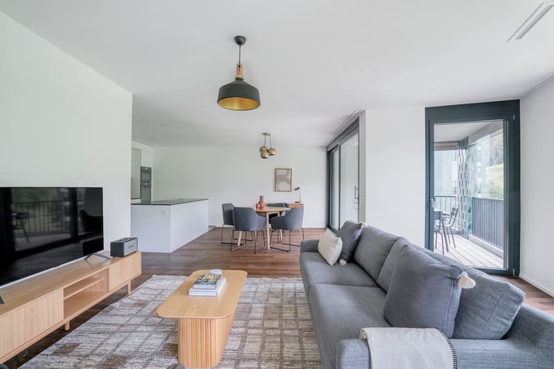 Open-concept living room with a large gray sofa, wooden coffee table, and a dining area with a wooden table and chairs. The room has a balcony or terrace with sliding glass doors, providing natural light and access to the outdoor space. The room features