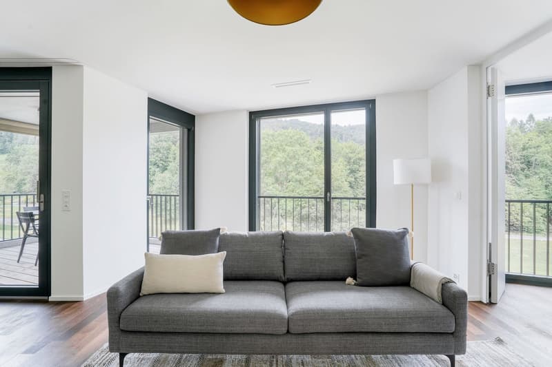 Spacious living room with large windows overlooking a forested area, hardwood floors, gray sofa, and a pendant light fixture.