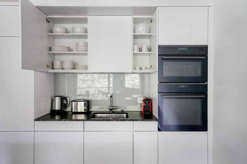 Modern, minimalist kitchen with white cabinets, open shelving displaying white dishes and bowls, black granite countertop, built-in oven and microwave, and a dishwasher