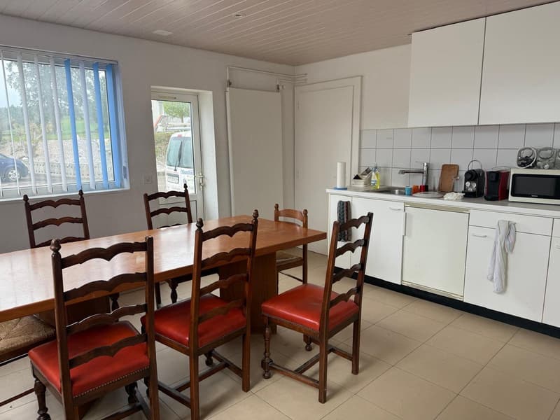 The image shows a combined kitchen and dining area. The kitchen features white cabinets and a tiled backsplash, with a dishwasher visible. The dining area has a wooden table with four ladder-back chairs with red cushions. The room has large windows provid
