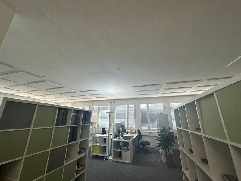 The image shows an office space with a desk, computer, and shelving units. The room has large windows, a suspended ceiling with recessed lighting, and a neutral color scheme of white and gray.