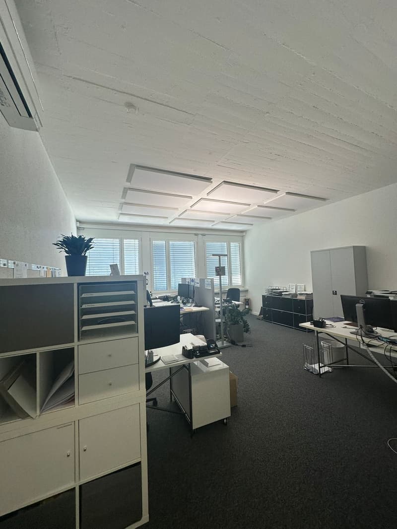 The image shows an office space with a raised ceiling, white walls, and a grid-like pattern on the ceiling. The room contains various office furniture and equipment, including filing cabinets, desks, and computers.