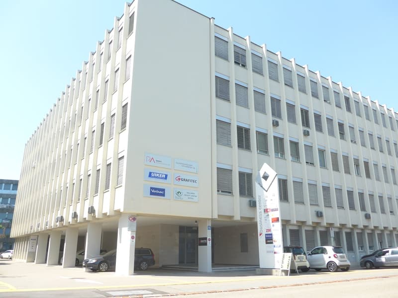 A multi-story office building with a modern, minimalist design. The building has a rectangular shape with rows of windows and ventilation systems. Several company logos are displayed on the facade, indicating that the building houses various businesses an