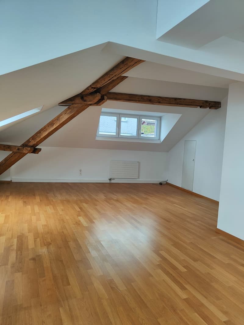 large open space with wooden flooring, wooden beams, white walls, a window, and a white door
