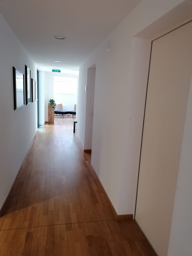 The image shows a long, well-lit hallway with white walls and a hardwood floor. There are several doors along the hallway, and at the end of the hallway, there is a room with a table and chairs, suggesting this is an office or workspace area.