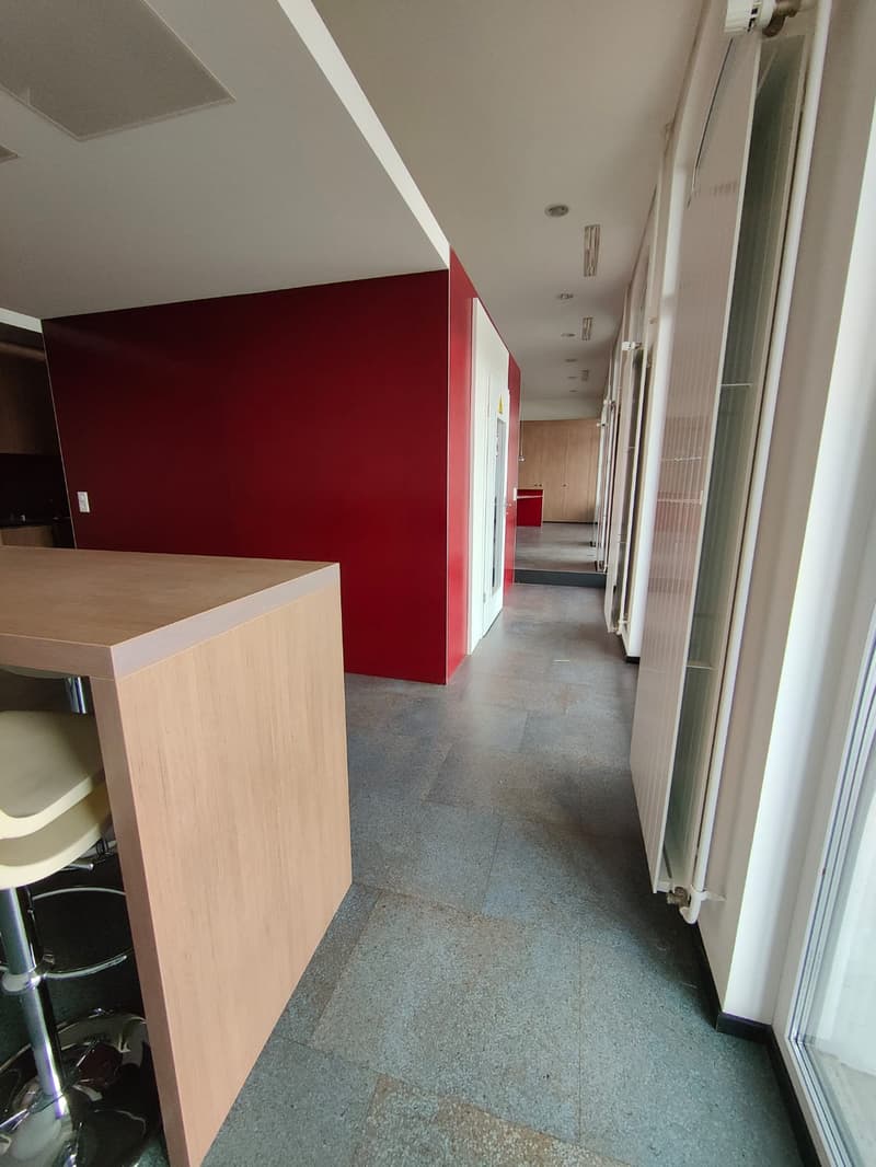Wide room with a counter table, a red wall, wooden floors, open hallways, and glass windows.