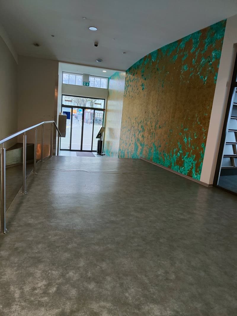 A large lobby with concrete flooring, a metal handrail on the stairs, a wall with green and brown paint, and glass doors leading to the outside with bicycles parked.