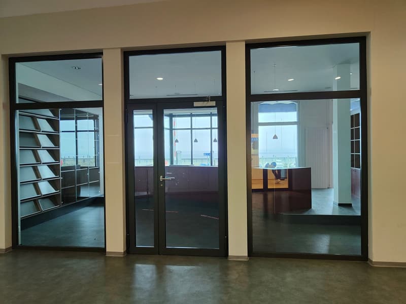 Large glass entrance doors, empty room, concrete floor, glass shelves on left, large window on right.