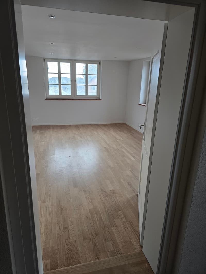 An empty room with white walls, hardwood floors, and a glass window letting in natural light. There is a white door to another room.
