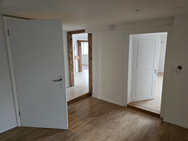 Open hallway with wooden floors, white doors, wooden beams, and an open door leading to another room.