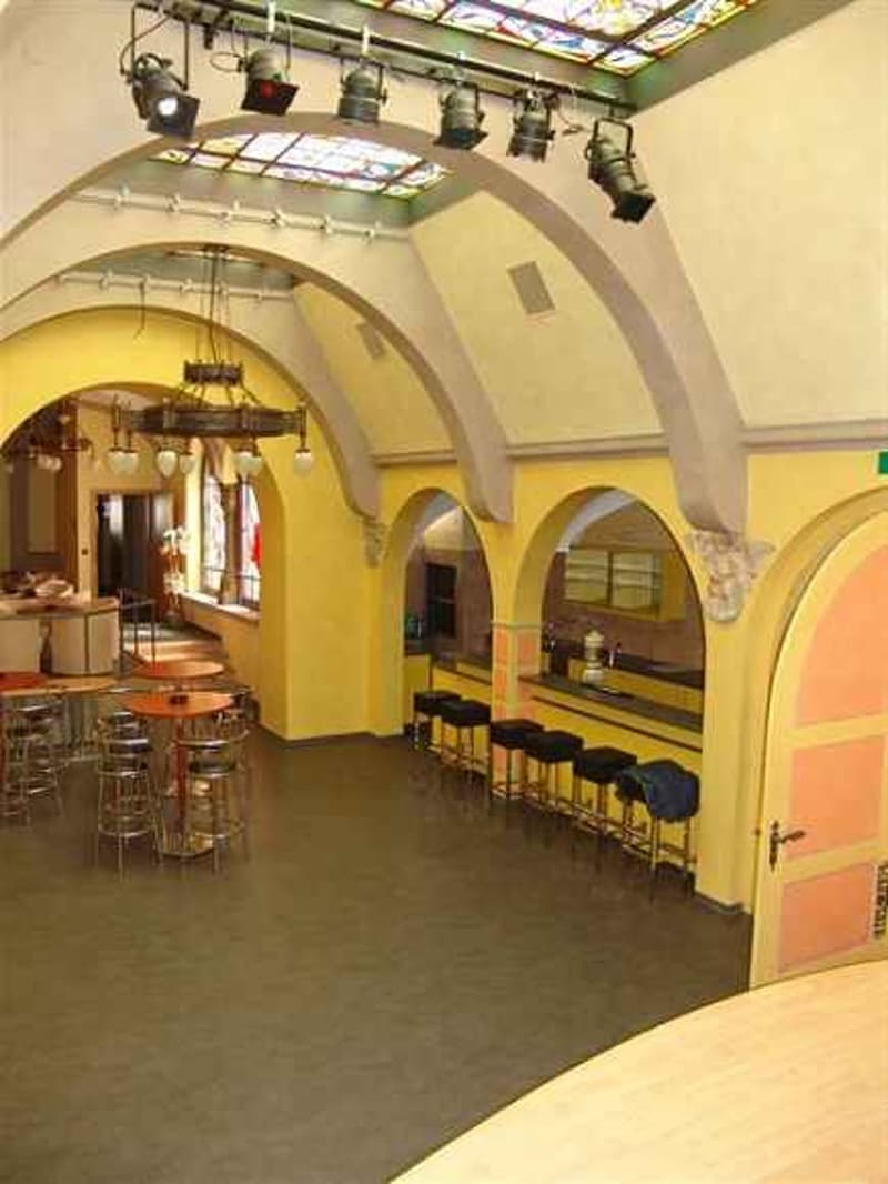 A large room with arched ceilings, yellow walls, and a wooden floor. There are tables and chairs, a chandelier, and a bar counter with stools. There are also lamps hanging from the ceiling.