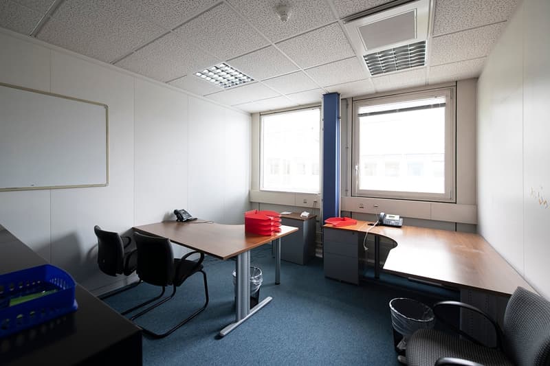 This image shows an office space with a desk, chairs, and storage units. The room has large windows providing natural light, and the ceiling has a grid of lighting fixtures. The floor is covered in blue carpeting.