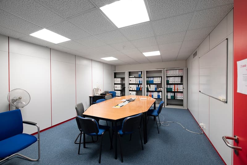This image shows an office space with a conference table, chairs, bookshelves, and storage cabinets. The room has a modern, professional appearance with gray walls and ceiling tiles. There is a fan visible, as well as a dishwasher, indicating this may be