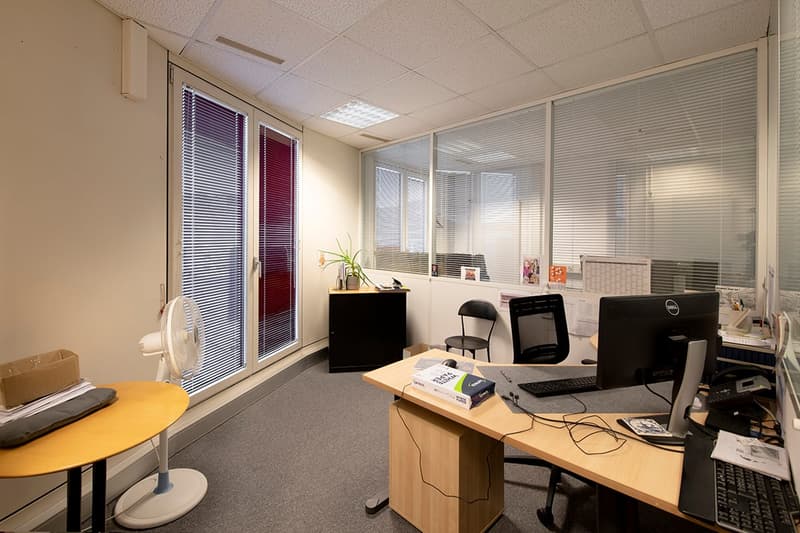 The image shows an office space with cubicles, desks, computers, and office equipment. The room has a neutral color scheme with beige walls and gray carpeting. There are blinds on the windows and overhead lighting fixtures. The office appears to be set up
