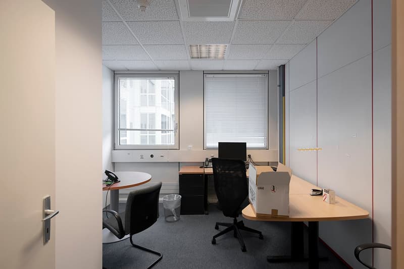 Small office space with a desk, chair, and computer. The room has a window, ceiling lights, and some storage boxes. The floor is carpeted.