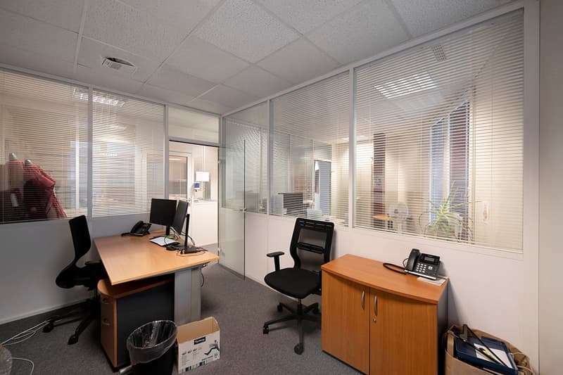 The image shows an office space with multiple workstations. The room has a suspended ceiling, glass partitions, and office furniture such as desks, chairs, and a cabinet. The space appears to be well-lit with natural light coming in through the windows.