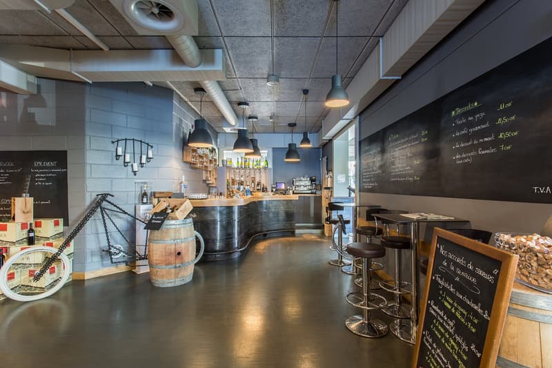 The image shows the interior of a restaurant or bar. It has a wooden bar counter with stools, shelves with bottles and glasses, and a chalkboard menu on the wall. The ceiling has exposed pipes and lighting fixtures, creating an industrial-style decor. The