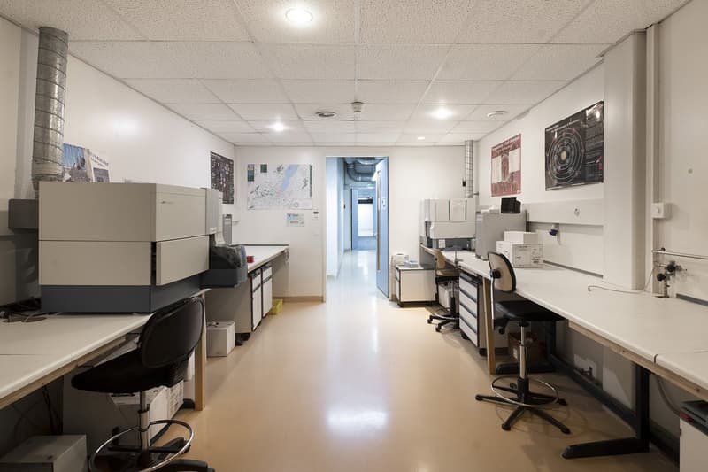 Industrial style laboratory with white walls, ceiling, and flooring, multiple workstations, adjustable chairs, various equipment, open corridor, light fixtures, bulletin boards with charts and posters, floor-mounted printer/scanner.