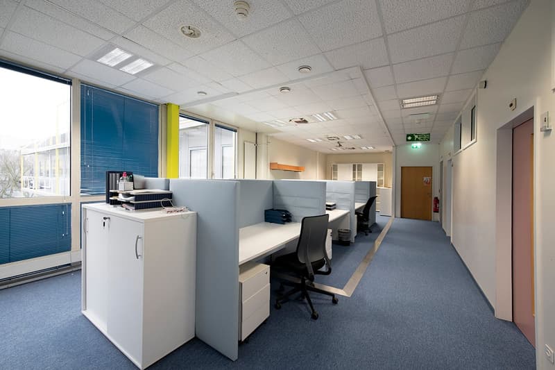Open office layout with cubicles, desks, and chairs. Bright lighting from windows and ceiling fixtures. Refrigerator and other office equipment visible. Hallway with doors leading to other rooms.