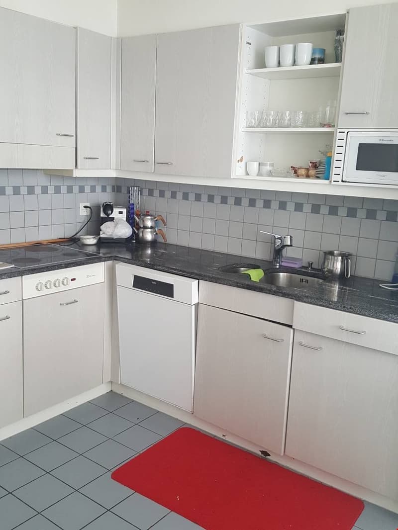 White cabinets, gray tile backsplash, black countertop, dishwasher, microwave, shelves with dishes and cups