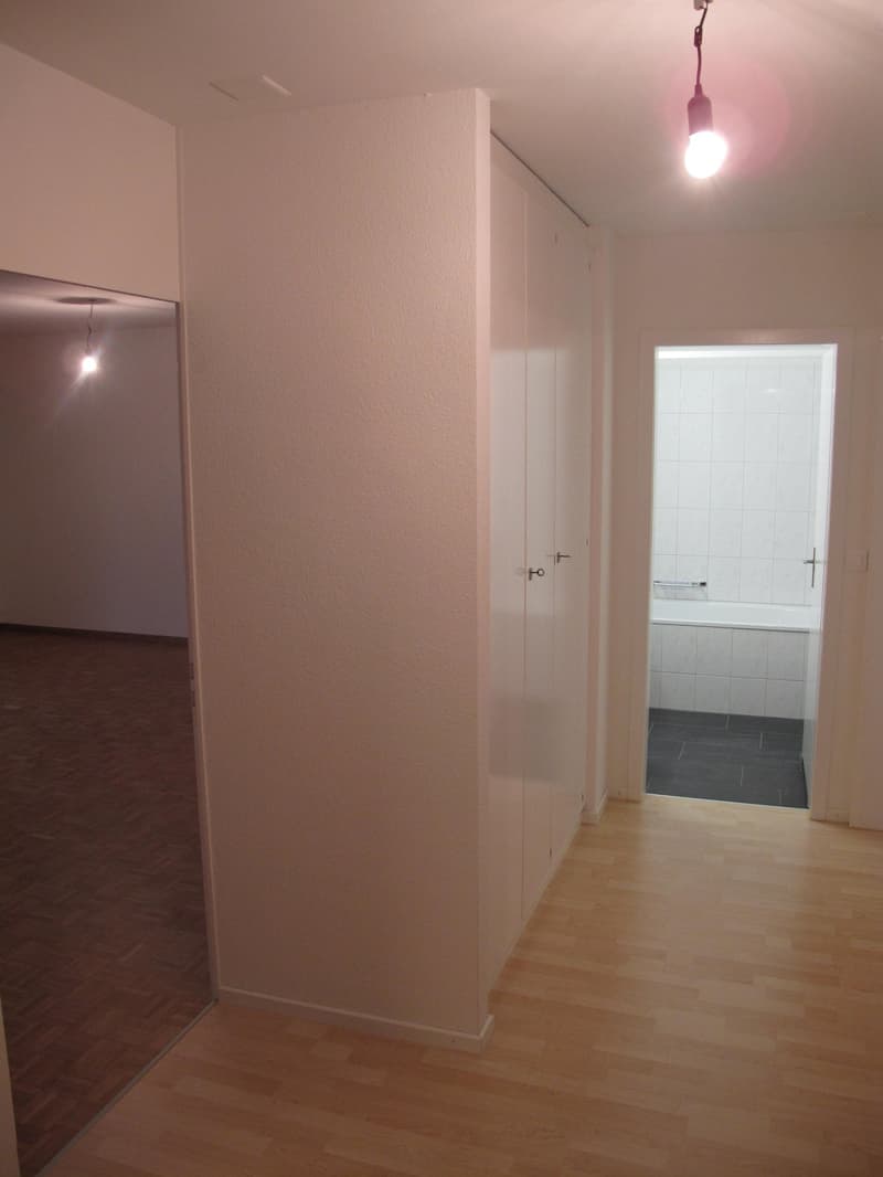 Empty room with wooden floor, white walls, two doors, one leading to a bathroom with bathtub, another leading to another room