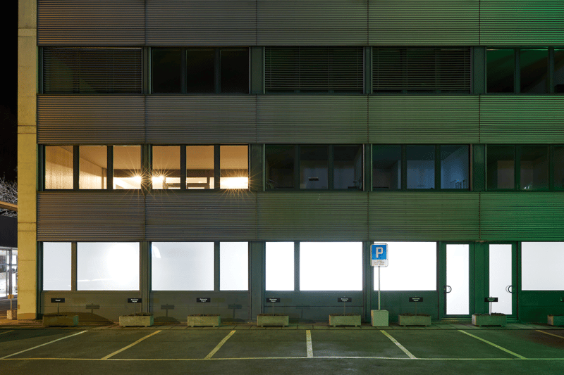 Modern commercial building, with several glass windows, green exterior, parking lot with several parking spots, sign for parking.