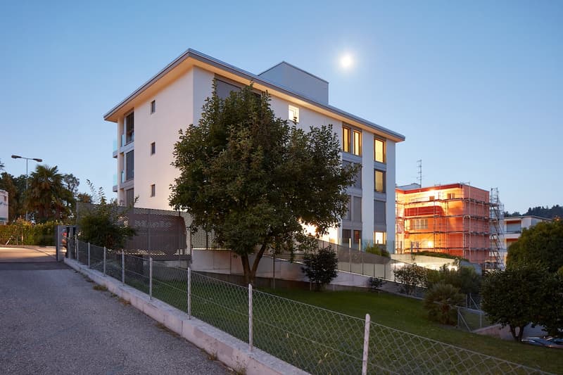 modern white building, multiple windows, fenced area, tree, street light, another building under construction