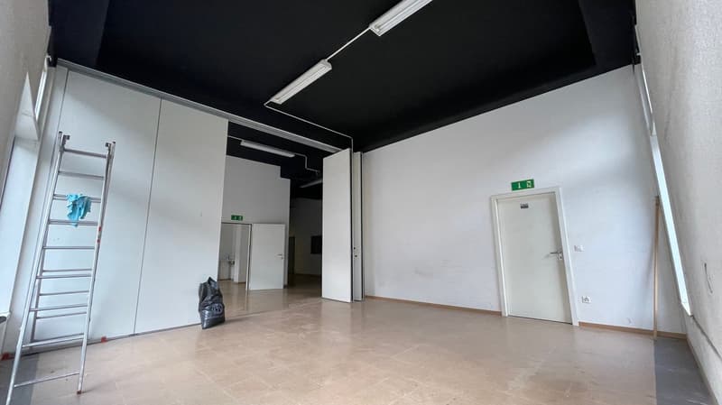 A large open space with a tile floor, white walls, a tall ceiling with light tubes, a white wall with a door on the right and a door in the middle.