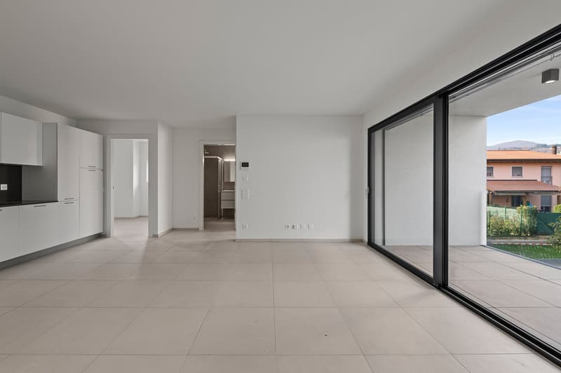 Spacious open-plan living area with large sliding glass doors leading to a balcony or terrace. The room has white walls, a modern kitchen with white cabinets, and a tiled floor throughout.