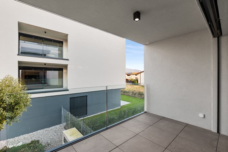 Modern apartment with large windows, glass balcony railing, and a view of the surrounding landscape and buildings.